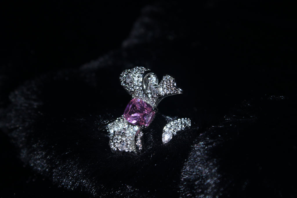 A silver-toned ring featuring a central pink gemstone and pavé-set clear stones, displayed on a black velvet surface.