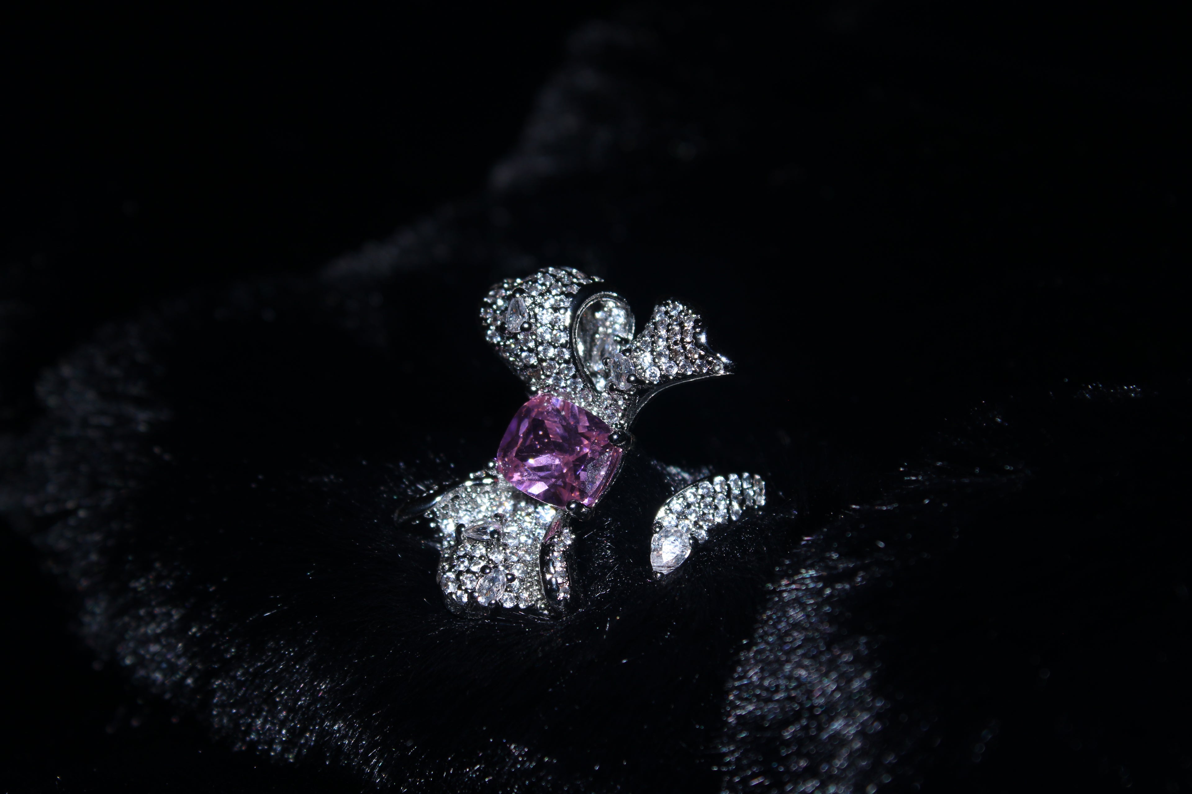 A silver-toned ring featuring a central pink gemstone and pavé-set clear stones, displayed on a black velvet surface.