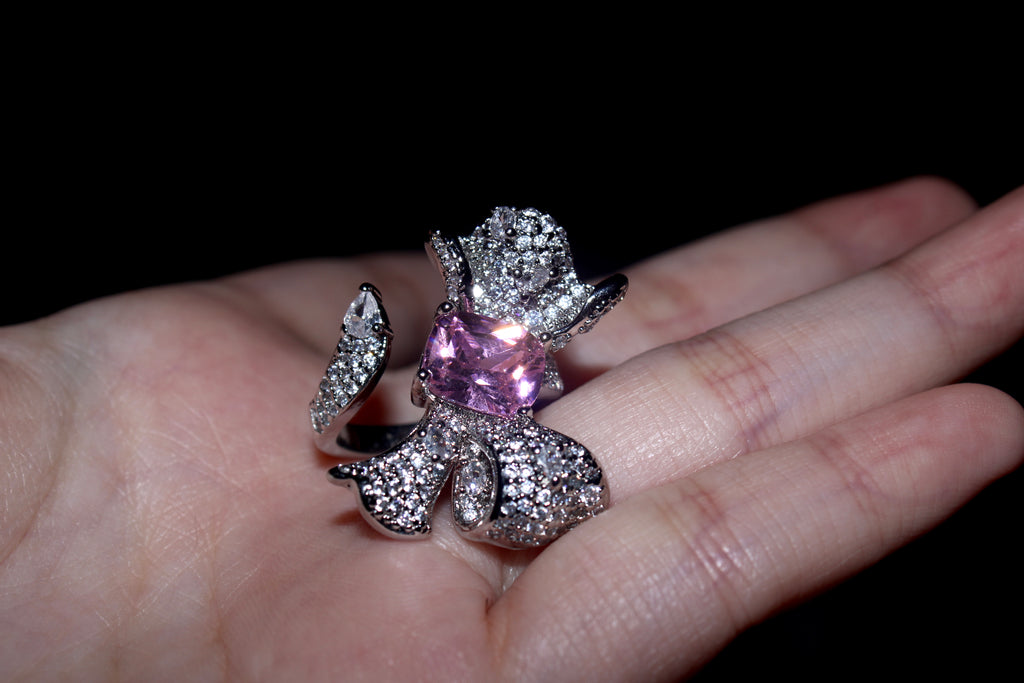 A silver-toned ring shaped like a flower, featuring a prominent pink gemstone in the center and pavé-set clear stones around it, shown resting on a person's palm against a black background.