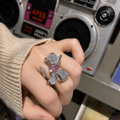 A close-up of a hand wearing a large, elaborate flower-shaped ring with a pink center stone and sparkling silver-toned petals, set against a blurred vintage boombox background.