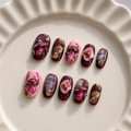 Ten decorative press-on nails arranged on a cream-colored ceramic plate. The nails feature glossy dark burgundy, pink, and marbled designs with raised floral and metallic gold details.