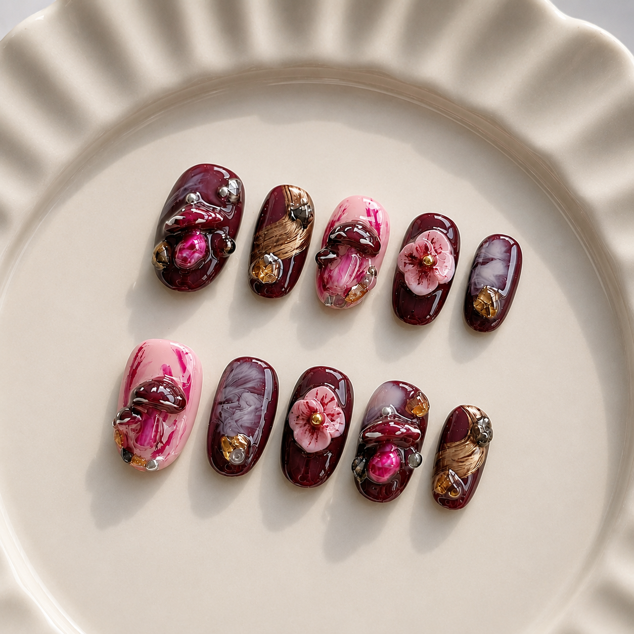 Ten decorative press-on nails arranged on a cream-colored ceramic plate. The nails feature glossy dark burgundy, pink, and marbled designs with raised floral and metallic gold details.